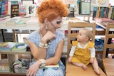A drag performer sits on a chair beside a small child