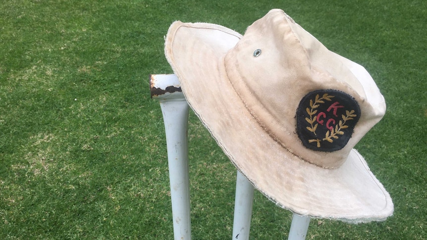 A well-worn cricket hat sits on a set of metal stumps on a pitch.