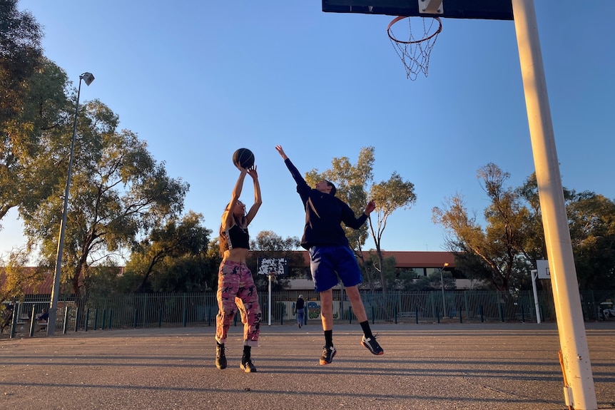 two people playing basketball, one shoots at goal, the other jumps up to defend