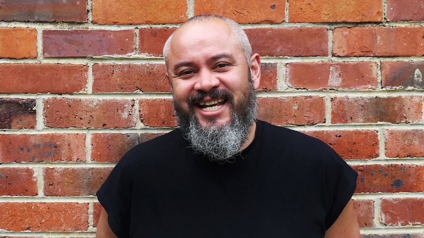 Balding man with salt-and-pepper beard, smiling, standing against red-brick wall.