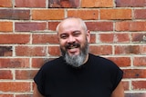 Balding man with salt-and-pepper beard, smiling, standing against red-brick wall.