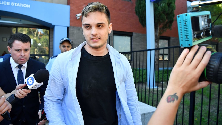 A man in a black t-shirt and blue shirt walks from a police station surrounded by media and cameras.