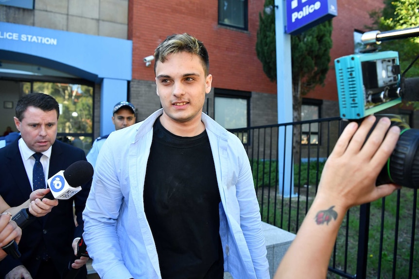 A man in a black t-shirt and blue shirt walks from a police station surrounded by media and cameras.