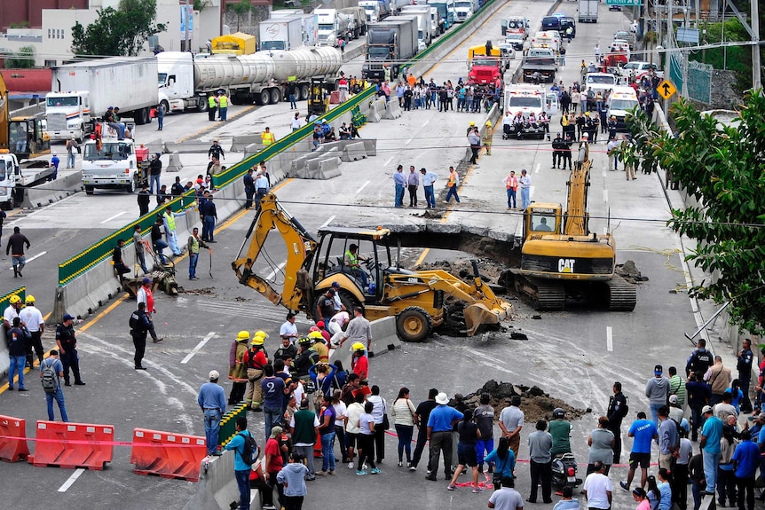A large group of people gather to watch rescue workers remove a car from as sinkhole, in the middle of a highway.