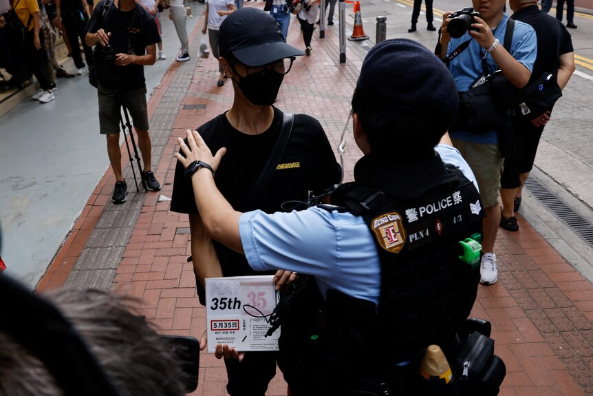 A police officer stands in front of a man who is dressed in black and holds a sign that says 'May 35'. 