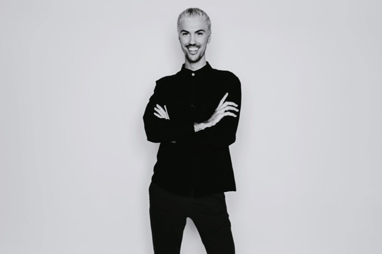 A black and white photo of a young man wearing all-black smiling at the camera, arms-crossed