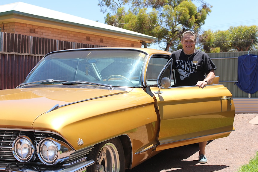 A man with brown hair and a black shirt standing inside the driver's-side door of a gold Cadillac.