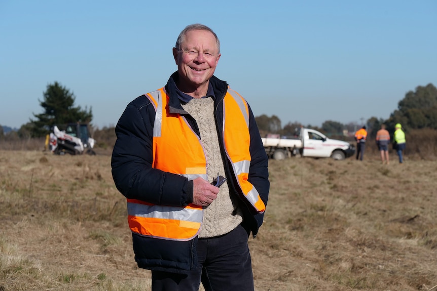 Ed Suttle stands in a largely empty paddock wearing a orange high vis vest and smiling.