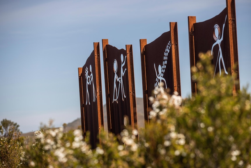 Rust coloured metal panels with Aboriginal artwork depicting the creation story, set among flowering native shrubs