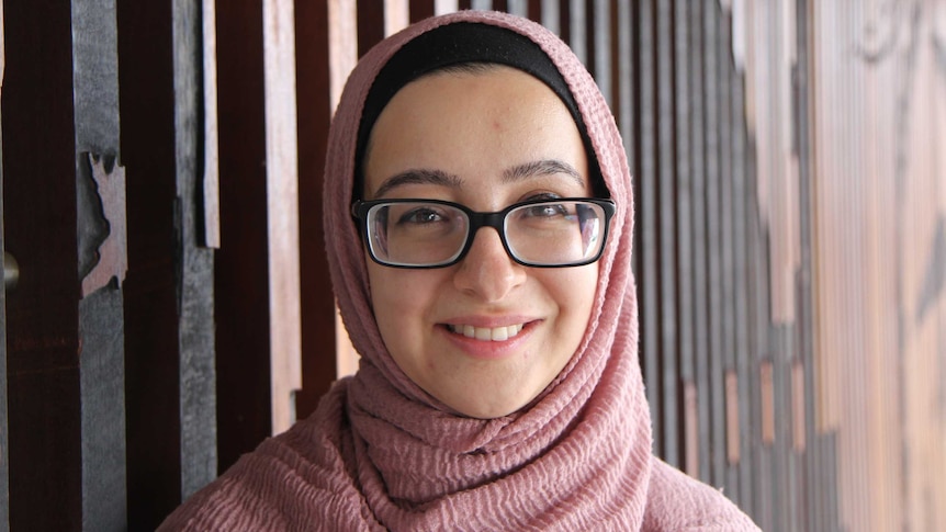 A woman in a pink headscarf stands in front of a decorative, wooden wall.