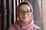 A woman in a pink headscarf stands in front of a decorative, wooden wall.