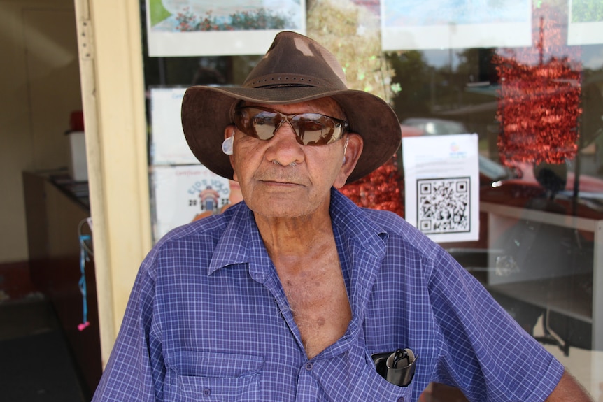 An indigenous man stands in a dark blue checkers shirt with brown sunglasses and a hat.