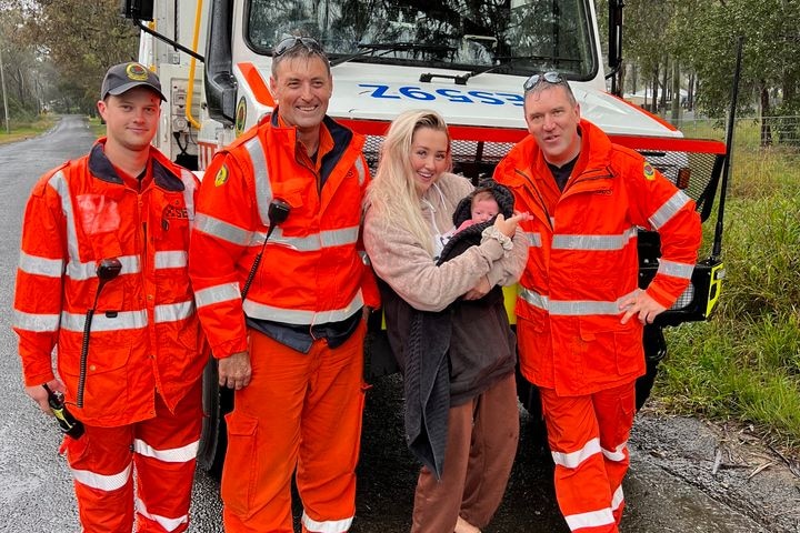 Two SES volunteers dressed in orange high-visibility suits stand on either side of a mother holding her baby.