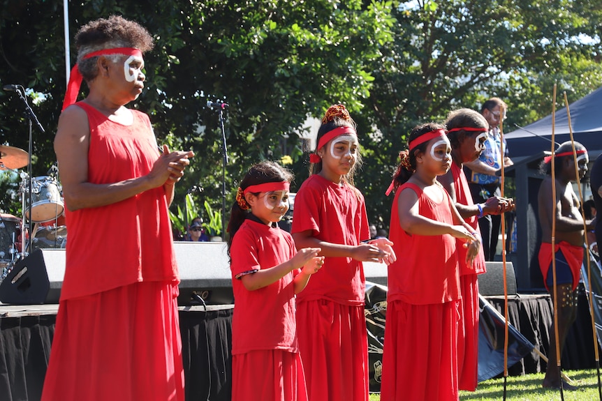 Young and older women wear red clothes and headbands with white face paint and dance near a stage