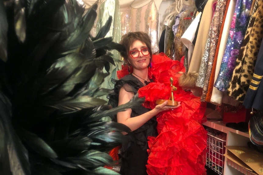 A woman with brown hair and bright red glasses stands in a costume wardrobe and holding a gold logie