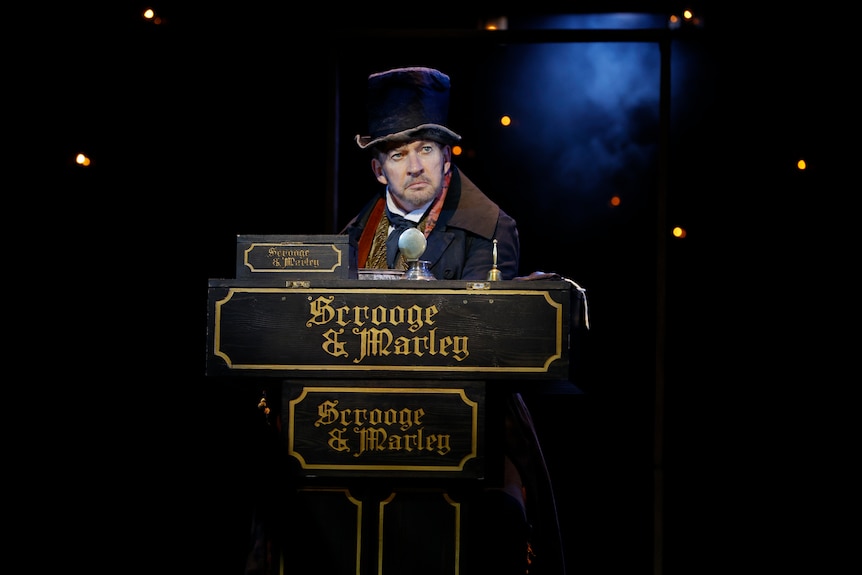 On stage, a middle-aged man in Victorian era dress looks disgruntled, standing behind a desk that reads "Scrooge & Marley"