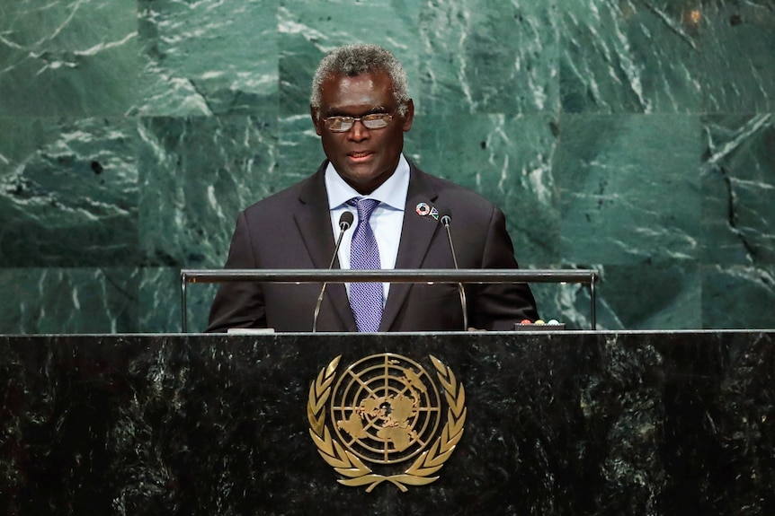 Man ina suit at a podium addresses a room full of people.