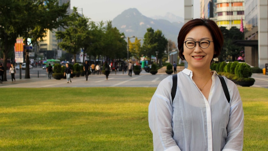Wideshot of a woman in glasses and white shirt with a cityscape in the background.