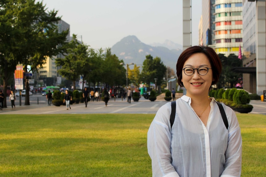 Wideshot of a woman in glasses and white shirt with a cityscape in the background.