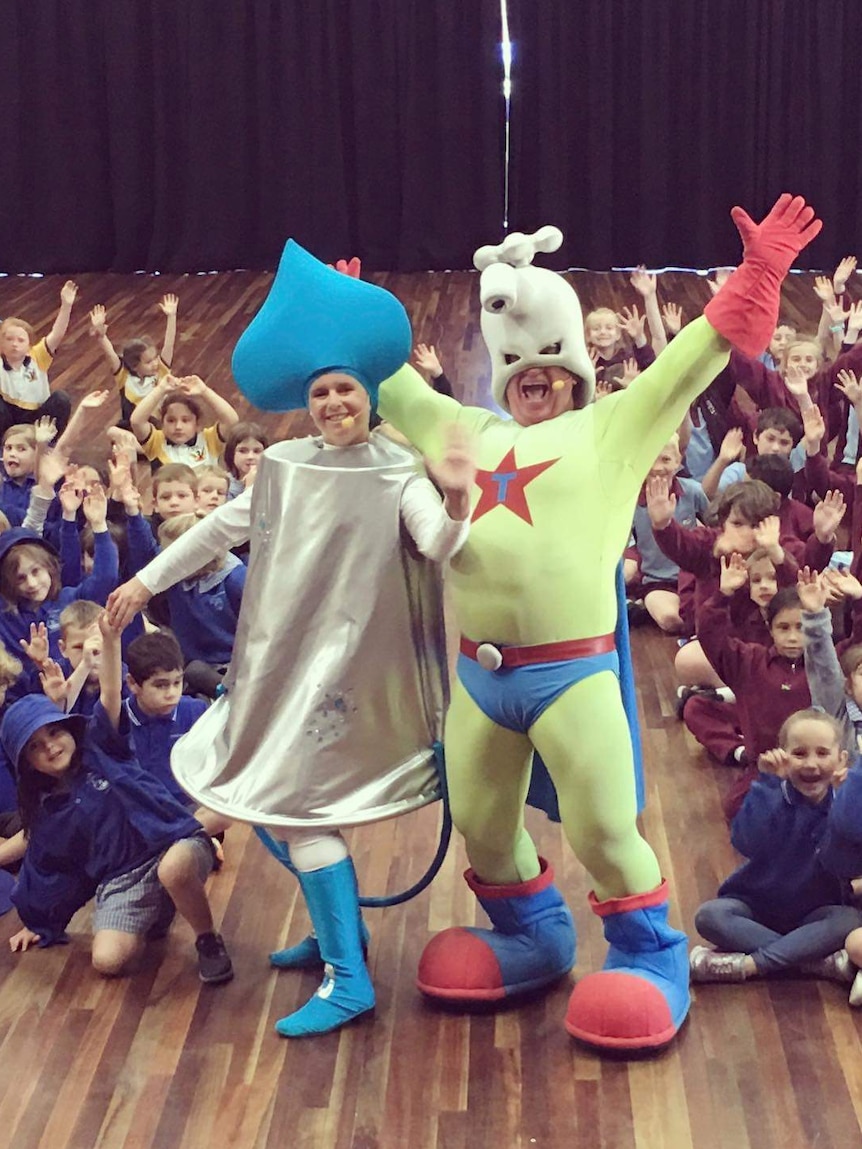 Two mascots are surrounded by small children in a school performance of their water awareness show