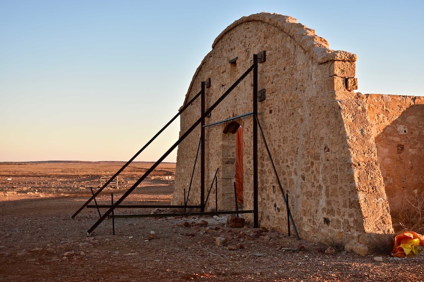 A large stone wall and archway is supported by a metal support against a blue sky at dusk.