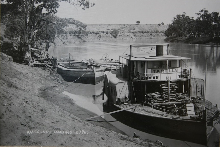 A black and white image of a small paddle steamer, it is tied up to a river bank next to a barge, an expansive river is behind