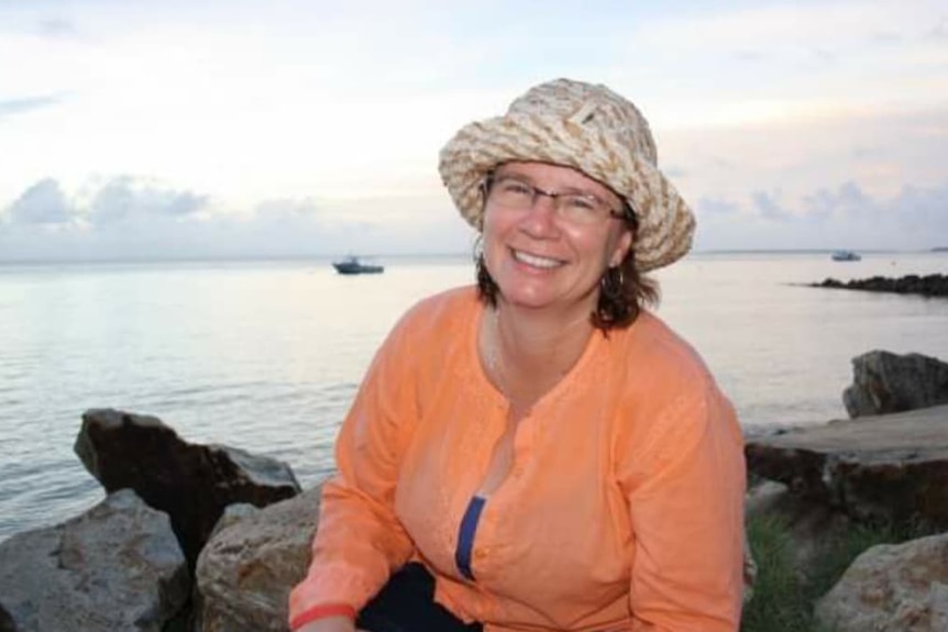 Michelle Van Dyk wearing an orange shirt and light coloured bucket hat sitting infront of the ocean at North Stradbroke Island