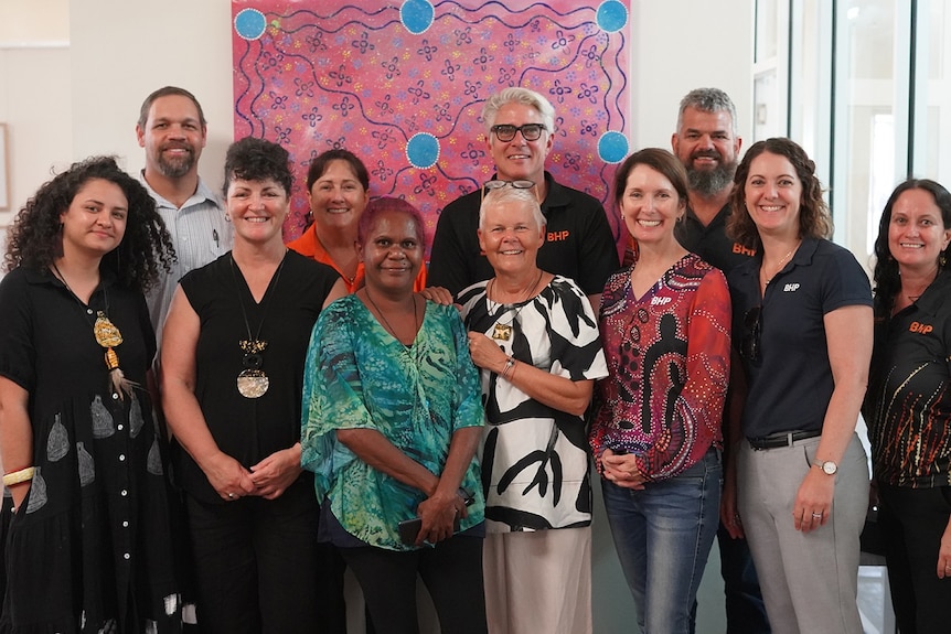11 people standing in front of a pink and blue Aboriginal artwork smiling at the camera.