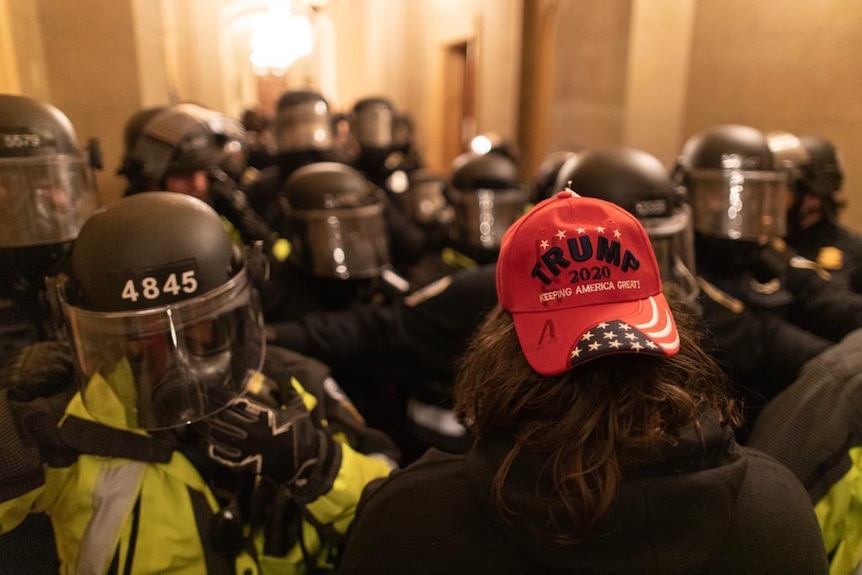 A man with long hair and a Trump 2020 hat faces armed riot police inside the Capitol building in Washington DC