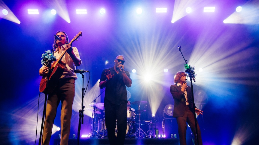 Idris Elba dressed in black, wearing sunglasses, shares the stage with Lime Cordiale (dressed in suits)