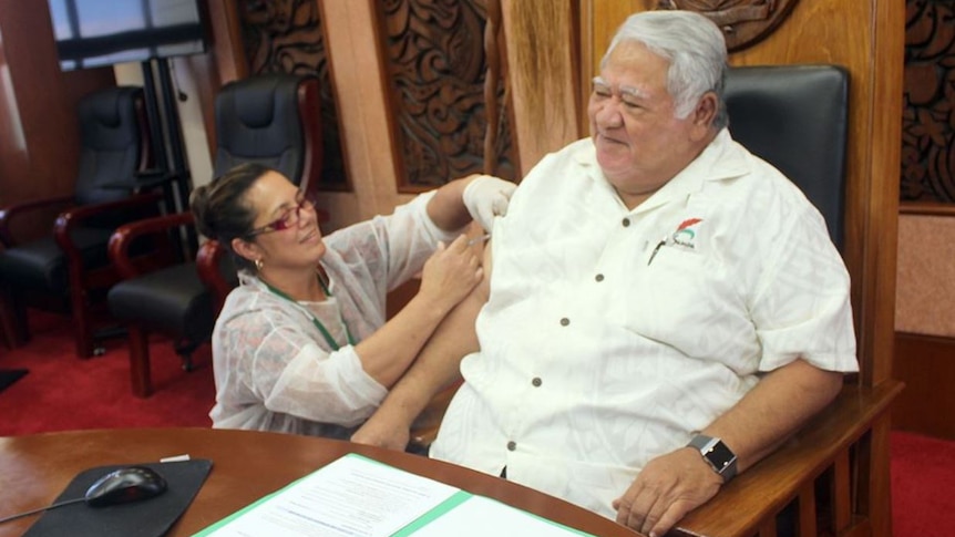 Samoan Prime Minister Tuilaepa Sailele sits on a large leather chair as a woman inserts a measles vaccination via needle.