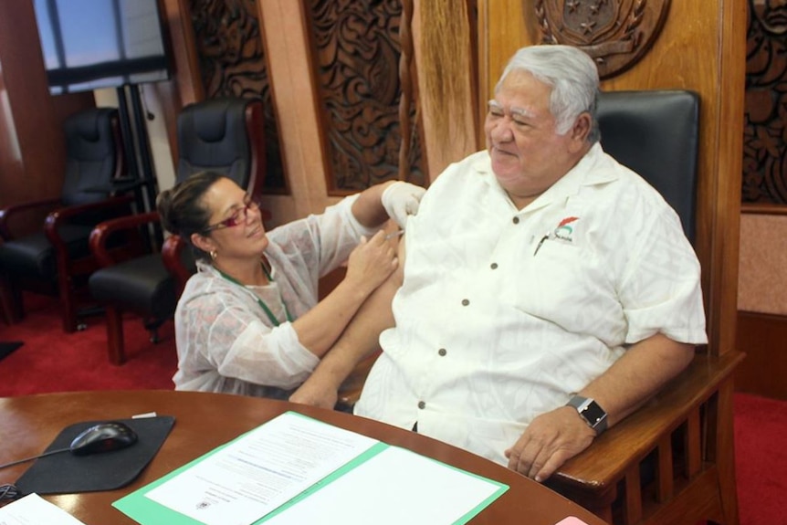 Samoan Prime Minister Tuilaepa Sailele sits on a large leather chair as a woman inserts a measles vaccination via needle.