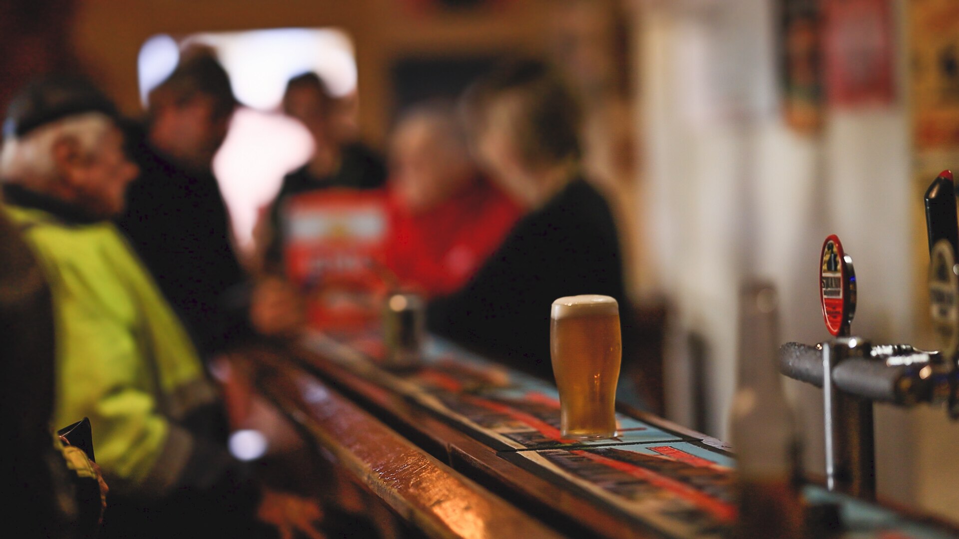 A pint of beer sits on a bar. Blurred people are in the background.