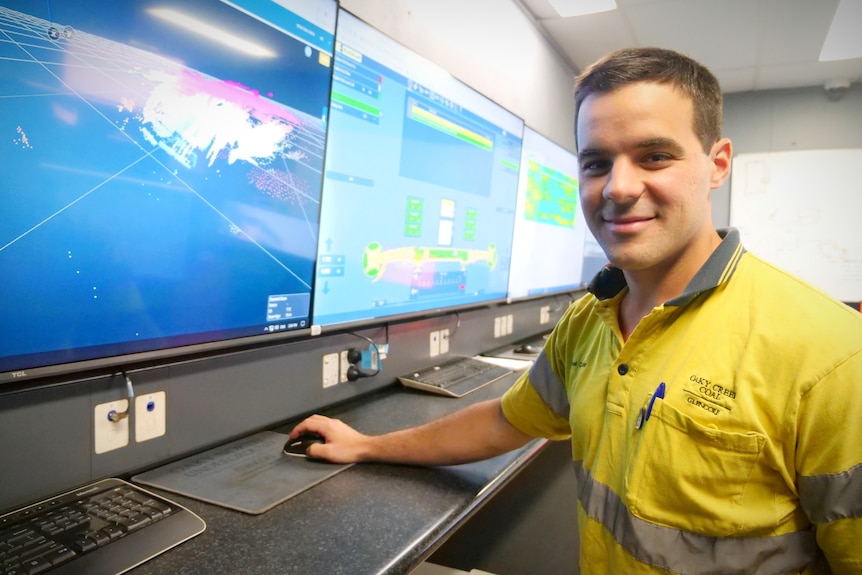 A man in a bold yellow shirt looks at a camera next to a wall of computer screens as he clicks a mouse.