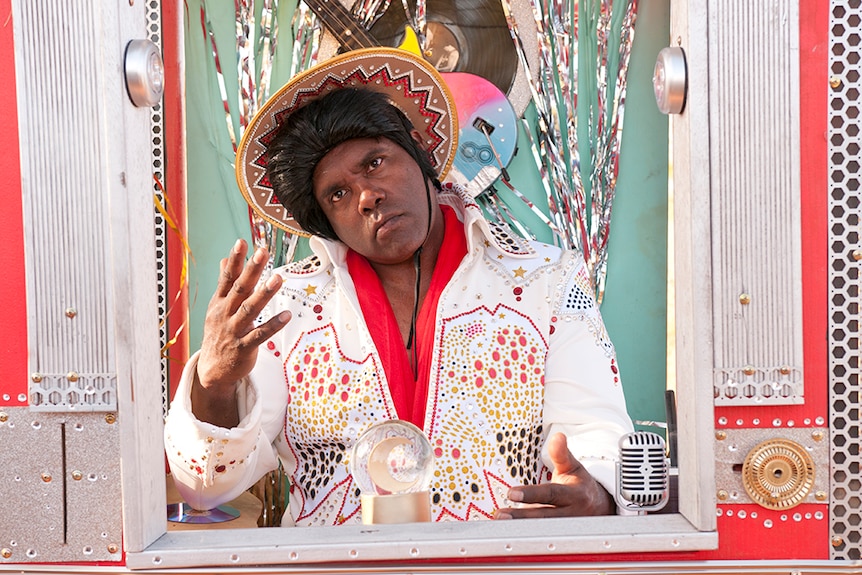 A man sits in front of streamers, bright coloured guitar and wears black wig and rhinestone decorated hat and jumpsuit.