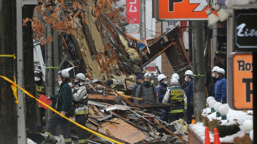 People in helmets stand among debris from buildings with snow visible