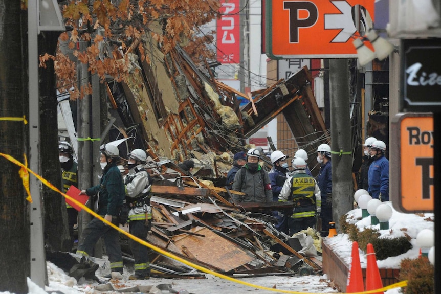 People in helmets stand among debris from buildings with snow visible