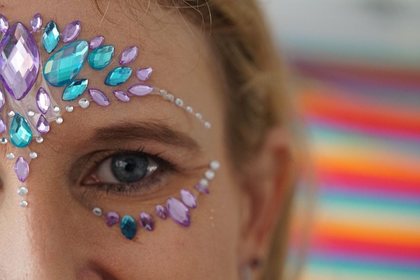 A close up of a woman's eye with glitter on her face and the pride flag in the background