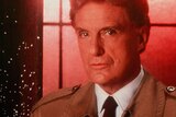 Robert Stack looks at the camera wearing a tie and trench coat, with a glowing red window behind him