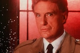 Robert Stack looks at the camera wearing a tie and trench coat, with a glowing red window behind him