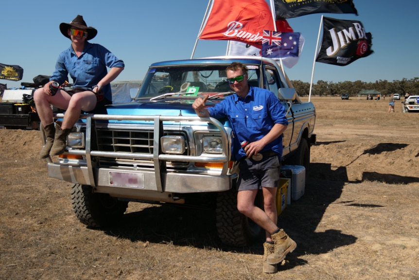 Two men in blue shirts, one in a black hat, pose next to a blue ute flying flags