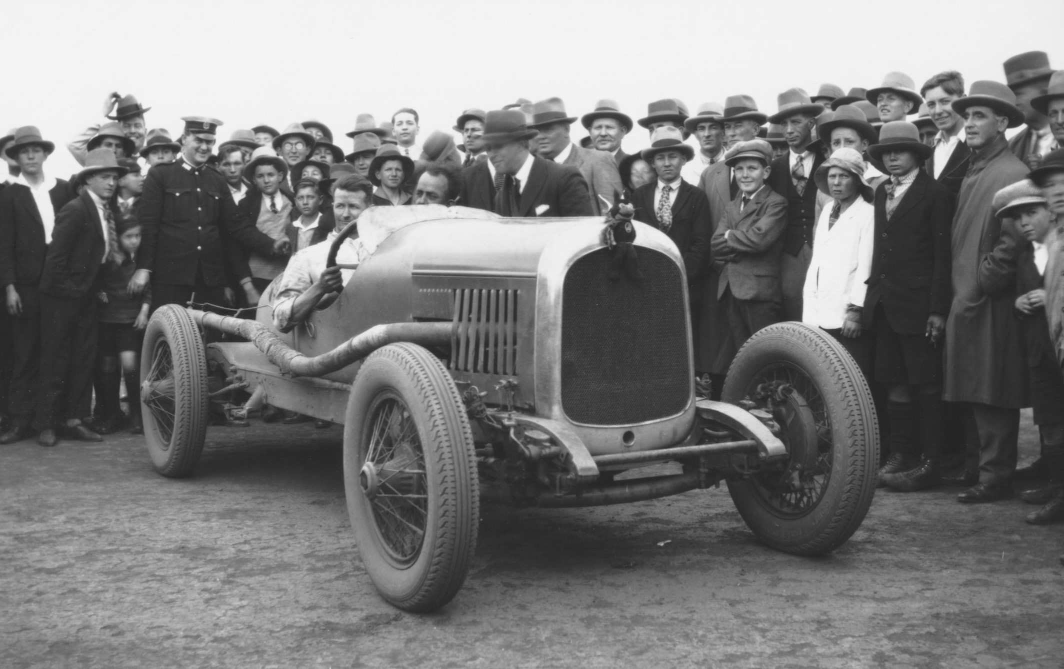 A black-and-white photo of a man sitting in a car surrounded by people.