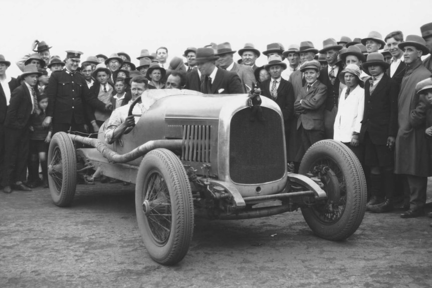 A black-and-white photo of a man sitting in a car surrounded by people.