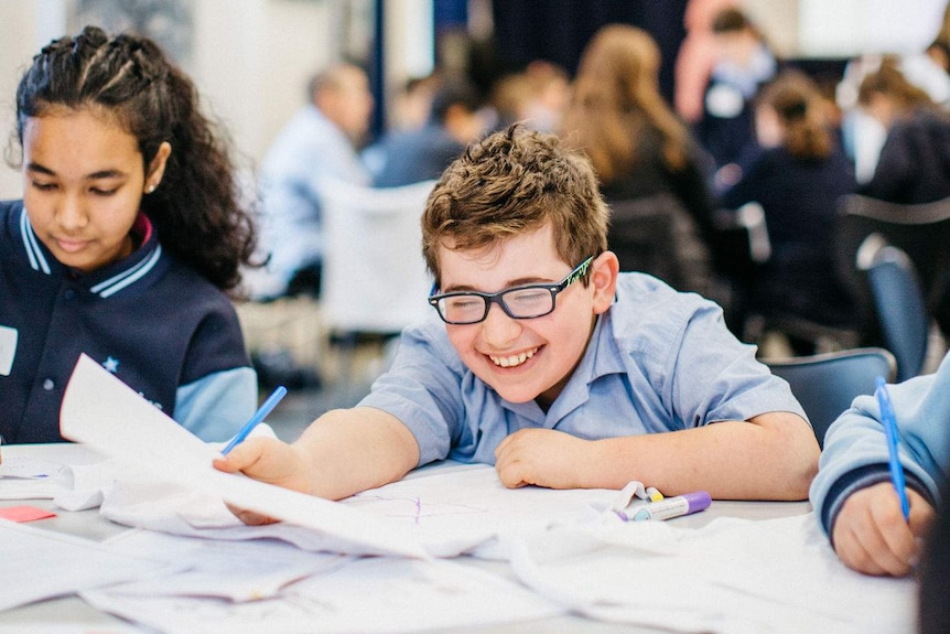 A young girl and boy in school uniforms sit at a table with coloured paper. The boy laughs, the girl has a focused expression.