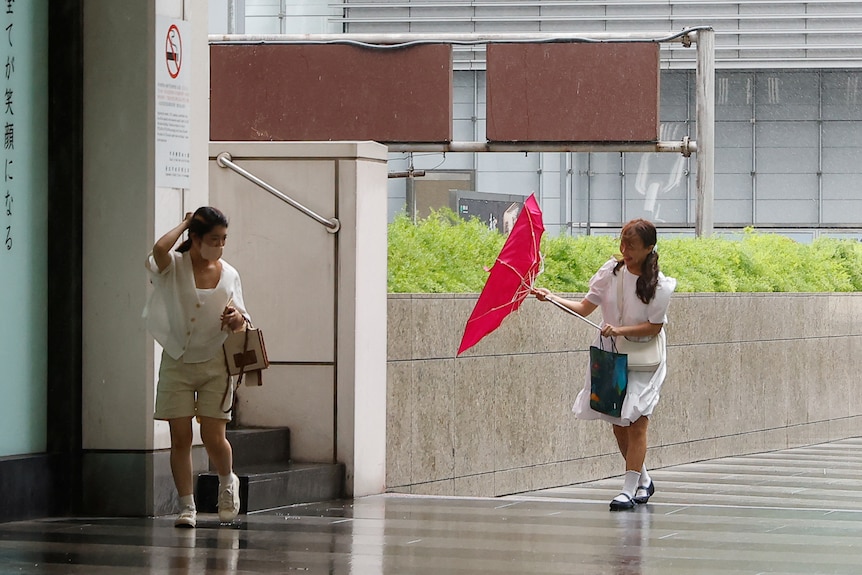 A woman holds onto her head and a second woman tries to fix her upside down umbrella
