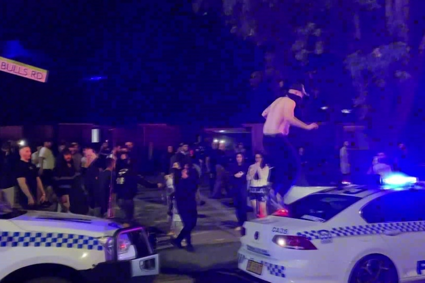 A person stands on top of a police car at night as a group of other people surround the car.