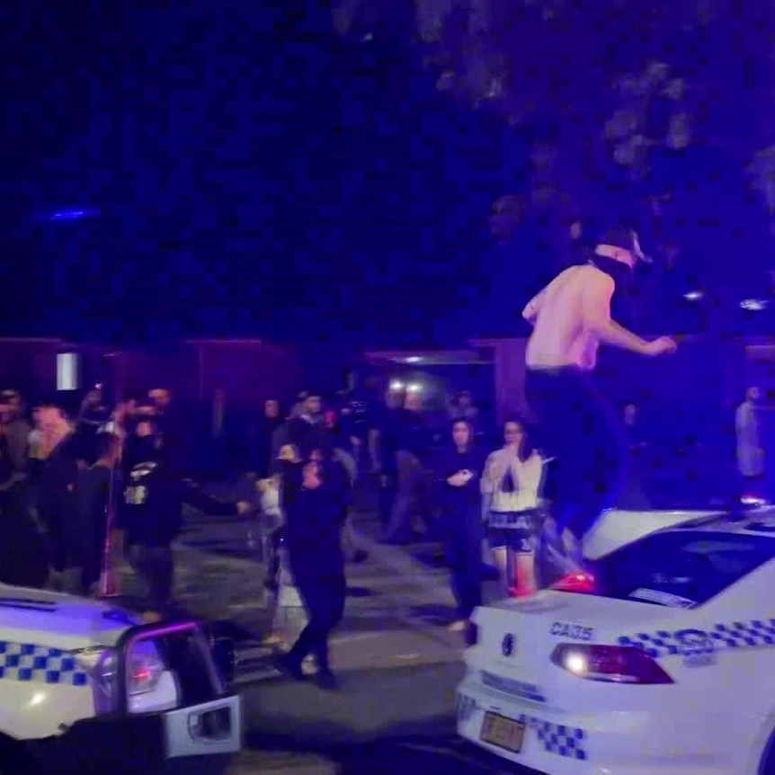 A person stands on top of a police car at night as a group of other people surround the car.