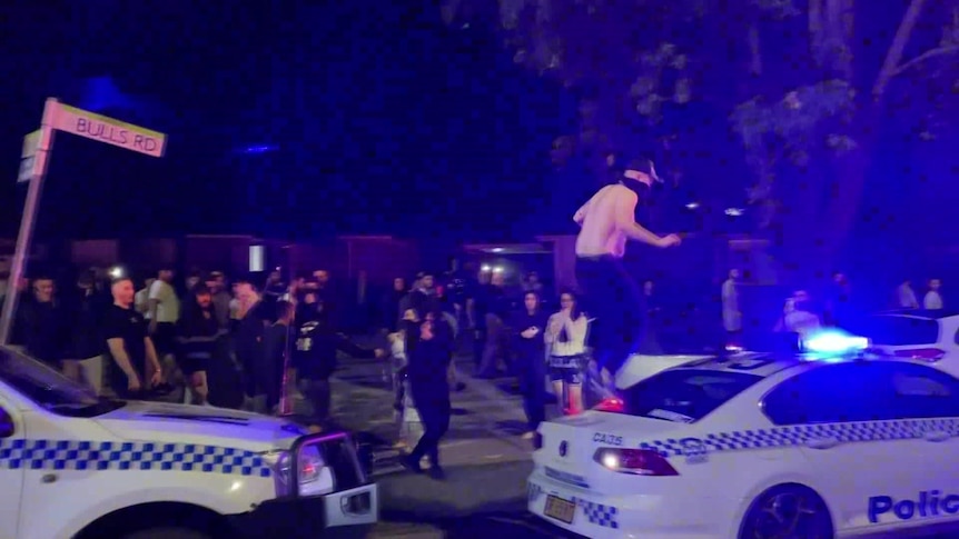 A person stands on top of a police car at night as a group of other people surround the car.