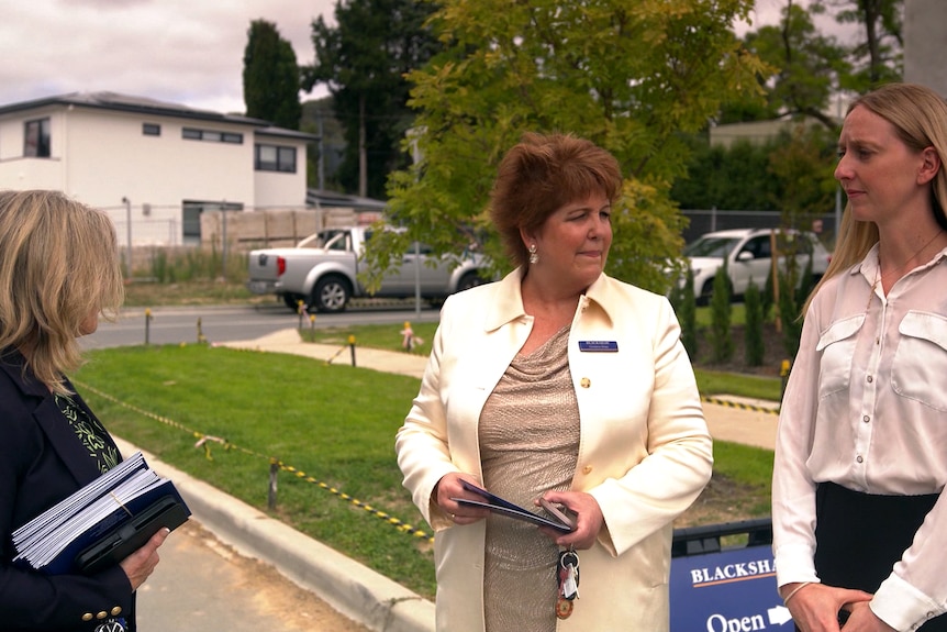 Christine Shaw, in a cream coat and with reddish-brown hair, stands in a street with 2 other people, holding a folder and keys.