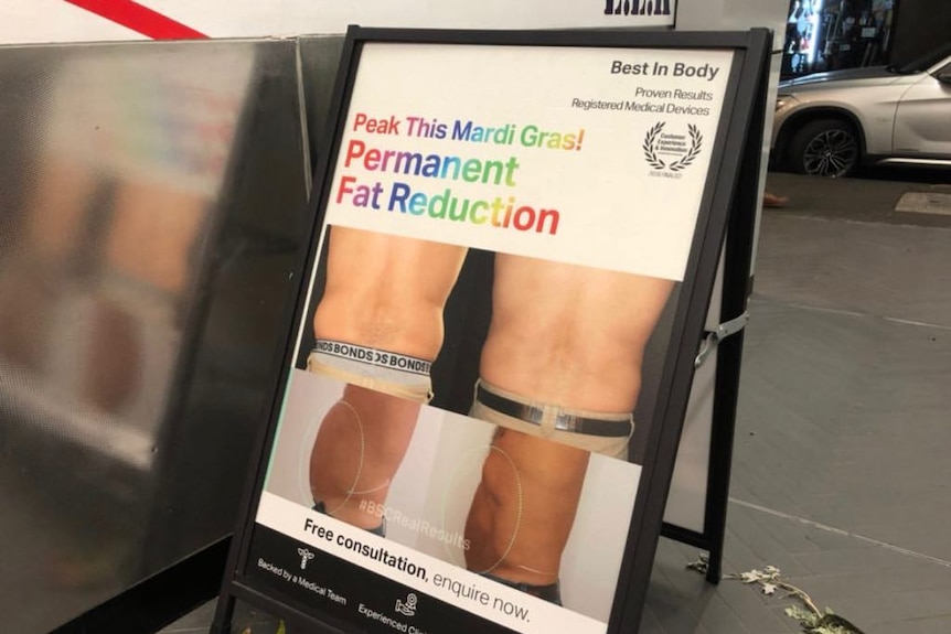A sandwich board that says 'Peak this Mardi Gras! Permanent fat reduction' on a street.